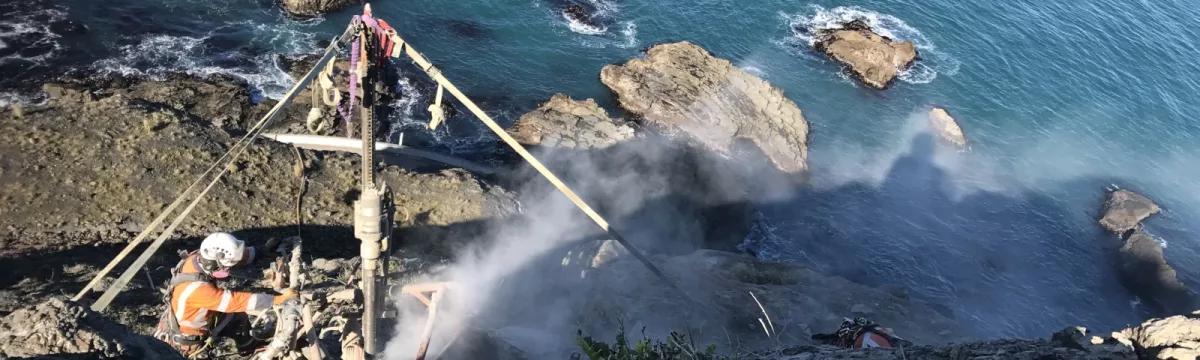 Avalon team member with a drilling rig on a rocky slope above the sea